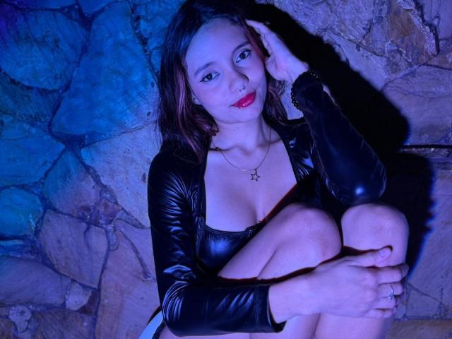 AmarandSofty, con la pelle chiara, il rossetto rosso e l'outfit in pelle nera, seduta con le gambe incrociate, una mano sulla testa, illuminata da una luce blu. sfondo con muro di pietra, collana con stelle, sexy, sguardo intenso, posa seducente.