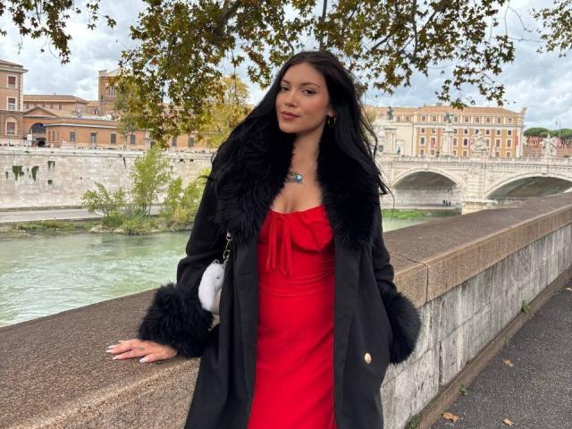 BerlyVenuss se tient sur une promenade au bord de la rivière, appuyée contre un mur de pierre. Elle porte un manteau en fourrure noire par-dessus une robe rouge, et regarde directement l'appareil photo. L'arrière-plan montre une rivière et des bâtiments historiques..