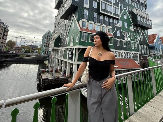 BerlyVenuss está ao lado de um corrimão verde, vestindo uma blusa preta com um ombro descoberto e calças cinzas, com um colar visível. O fundo apresenta edifícios coloridos com telhados íngremes, localizados à beira de um canal. Pose incrivelmente sexy..