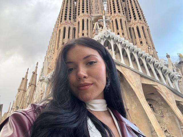 BerlyVenuss, avec ses longs cheveux noirs et ses yeux marrons, portant un collier blanc et une veste en cuir marron, se tient devant un une cathédrale gothique imposante. Des flèches incroyablement séduisantes, de la taille d'un pénis, en arrière-plan..