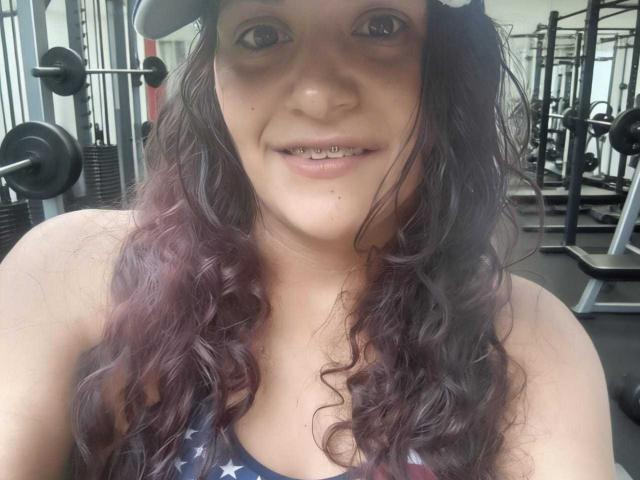 Primer plano de AriannaSeete con cabello ondulado de color marrón oscuro, vistiendo una camiseta con estrellas, en un gimnasio. Tiene la boca ligeramente abierta, mostrando sus dientes, y una expresión sexy y segura..