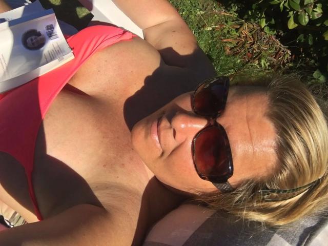 Close-up of Kim-Van-Dyke, a blonde woman with sunglasses, wearing a red bikini top, sunlight highlighting her cleavage, shadowed face, green foliage in the background, relaxed outdoor setting.