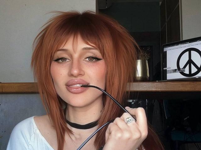 Fotografia de PhantomChristy, uma mulher de pele clara e cabelos ruivos, mordendo uma caneta de delineador preto, usando um colar preto. e uma blusa branca, com um visual sedutor, com um símbolo de paz em um monitor..