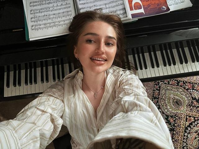 PhantomChristy, eine süße Frau mit heller Haut und braunen Haaren, lächelt, während sie ein Selfie vor einem Klavier macht. Sie trägt ein gestreiftes Hemd, über ihr liegen Notenblätter. Verdammt heiß..