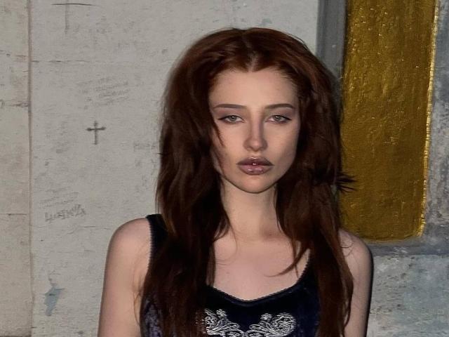 Fotografia di PhantomChristy con lunghi capelli castano scuro, che indossa un top nero in pizzo. Ha un'espressione neutra, le labbra leggermente dischiuse e una corporatura esile e pallida. Sfondo con texture dorata.