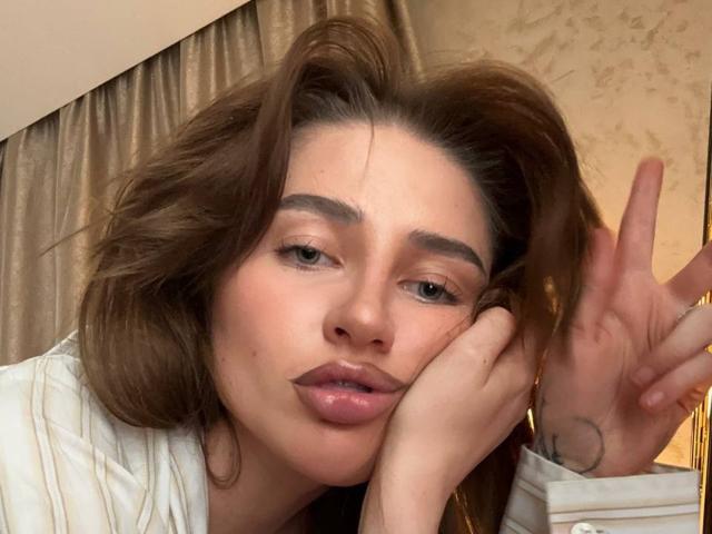 Close-up of PhantomChristy, a young woman with tousled brown hair, full lips, and dark eyebrows. She's making a peace sign with her right hand, looking seductive, with a slightly flushed face.