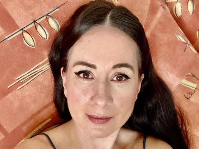 Close-up de heisseVicki-hot, uma mulher com cabelos longos e pretos, pele clara e olhos castanhos, deitada em uma superfície laranja. Luzes decorativas em forma de cordão ao fundo. Ela tem um sorriso discreto..