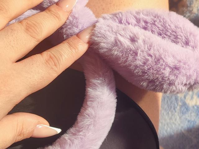 Primer plano de la mano de MinistraX, con uñas largas y cuidadas, sosteniendo una toalla suave y morada. La toalla cubre un pene grande y erecto. La luz solar brillante resalta la textura de la toalla y la piel..