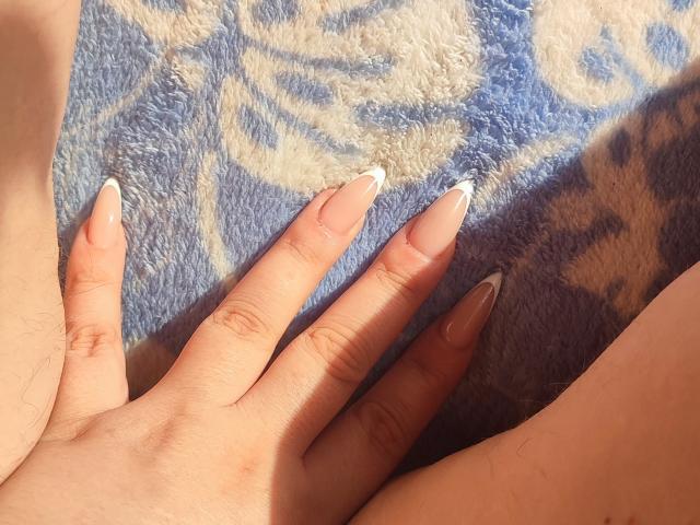 De hand van MinistraX, met lange, verzorgde nagels, rust op een deken met een blauw en beige patroon. Het zonlicht accentueert haar lichtbruine huid. Haar vingers zijn licht gespreid, wat een gevoel van ontspanning suggereert. Zachte texturen.