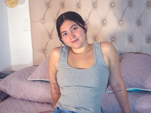 Mia Monroe è seduta su un letto, indossando una canottiera grigia aderente che mette in mostra il suo seno piccolo. Ha dei tatuaggi sulle braccia, i capelli scuri e un leggero sorriso. Sfondo: testiera imbottita..