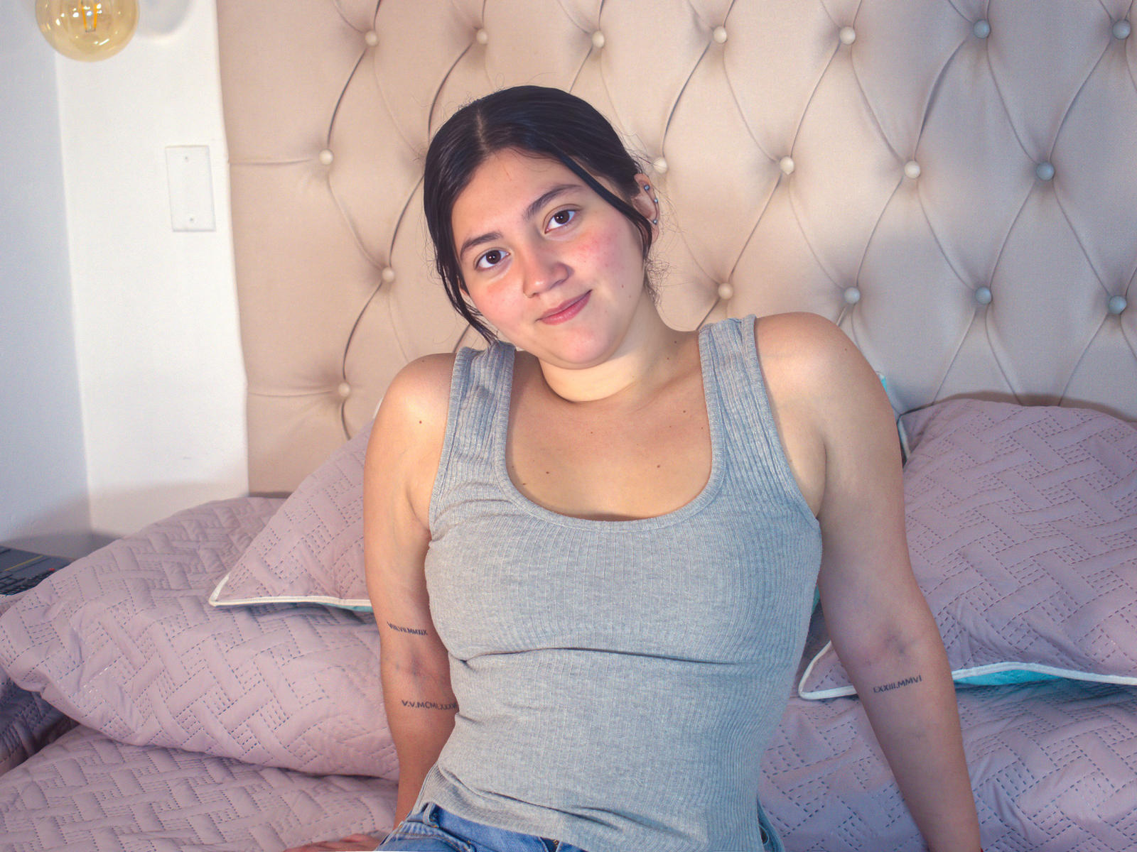Mia Monroe está sentada em uma cama, vestindo uma camiseta cinza justa que expõe seus pequenos seios. Ela tem tatuagens nos braços, cabelo escuro e um sorriso discreto. Fundo: cabeceira com detalhes em relevo..