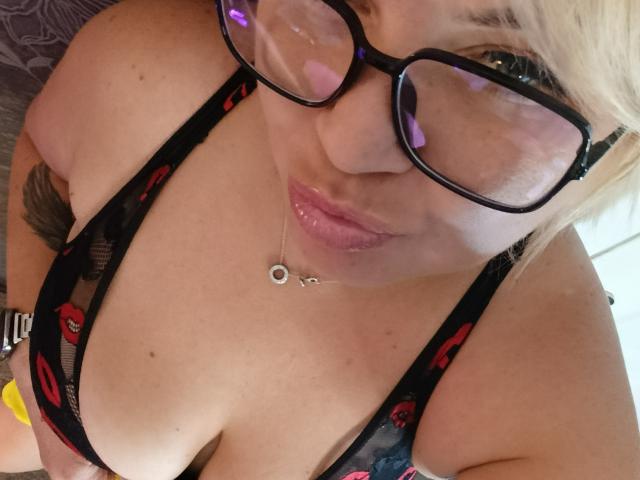 Gros plan de CurlyDreams portant des lunettes, avec des lèvres roses et un soutien-gorge en dentelle noire avec des motifs rouges. CurlyDreams a un collier avec un petit pendentif circulaire. Décolleté visible. Tension sexuelle.