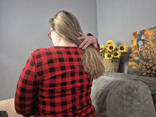 Viesta, mit dem Rücken zur Kamera, blonde Haare im Pferdeschwanz, rotes und schwarzes Karomuster-Hemd, Hand berührt das Haar, helles Sonnenlicht, graues Sofa. Sonnenblumen in einem Korb, farbenfrohes abstraktes Gemälde, lässig, sexuelle Spannung, intimer Moment..