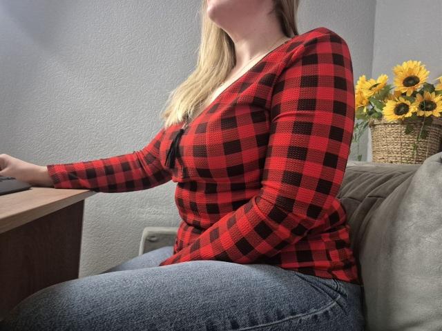 Viesta sits on a gray couch, wearing a red and black plaid shirt and blue jeans. Her blonde hair falls over her shoulder. Sunflowers in a basket behind her. Fucking tense vibe.