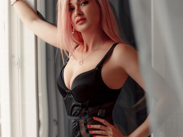 Photograph of Lustarela with pink hair, wearing a black, low-cut dress, standing in front of a window. She's pressing her hand against the window, looking seductively at the camera. Her red nails match her lipstick.