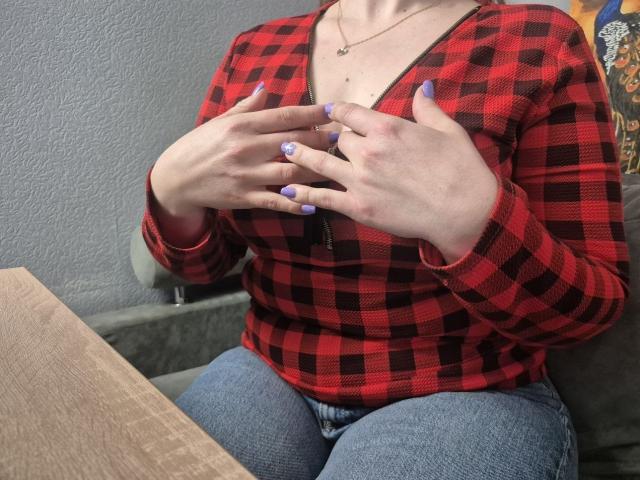 Viesta indossa una camicia a quadri rossa e nera, con le mani che stringono il petto, mostrando un seno di medie dimensioni. Unghie viola, jeans blu e una collana. Tensione sessuale con particolare attenzione al petto e alle mani..