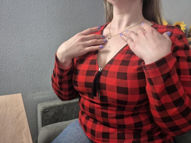Viesta, con una figura formosa e seno prosperoso, indossa una camicia a quadretti rossa e nera, con le mani che stringono il colletto, rivelando... un accenno di scollatura, smalto per unghie blu, parete grigia sullo sfondo.