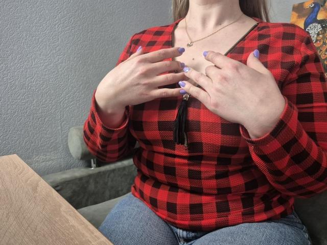 Viesta veste uma camisa xadrez vermelha e preta, com as mãos apertando o peito, com unhas pintadas de roxo, calças jeans azuis, de forma discreta. decote, colar com franjas, parede cinza, mesa de madeira, casual, sexy, tensão.