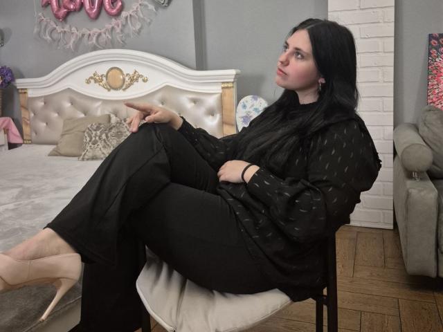 Esfil, avec sa peau pâle et ses cheveux noirs, est assise sur une chaise blanche, les jambes croisées, vêtue d'une robe noire et de talons beiges. Son expression est détachée, ses yeux sont tournés vers le haut. L'arrière-plan comprend une tête de lit capitonnée et des décorations en forme de cœur..