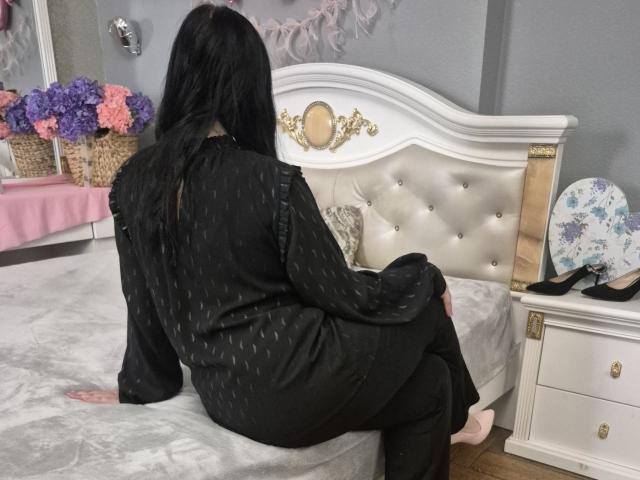 Una mujer con cabello negro largo, sentada en una cama blanca con cabecera acolchada, de espaldas a la cámara, vestida con una bata negra con lunares, con las piernas... ligeramente extendido, tensión sexual, decoración floral en el lado izquierdo.