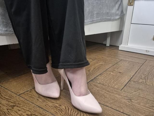 Les pieds d'Esfil, chaussés de talons aiguilles rose pâle, se tiennent sur un plancher en bois, son pantalon noir étant légèrement froissé autour des chevilles. Tiroir blanc avec une poignée dorée en arrière-plan. Élégance décontractée, incroyablement séduisante..