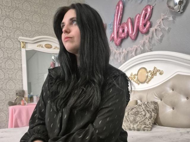 Esfil with long black hair sits on a bed, wearing a black coat, looking to the side with a slight smile. Pink "love" balloon in background. Soft, romantic lighting.