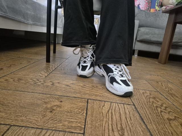 Esfil è in piedi, indossa pantaloni neri e scarpe da ginnastica bianche e nere con lacci bianchi, su un pavimento di legno. Un divano grigio con cuscini colorati è sullo sfondo. Un'atmosfera davvero rilassata..