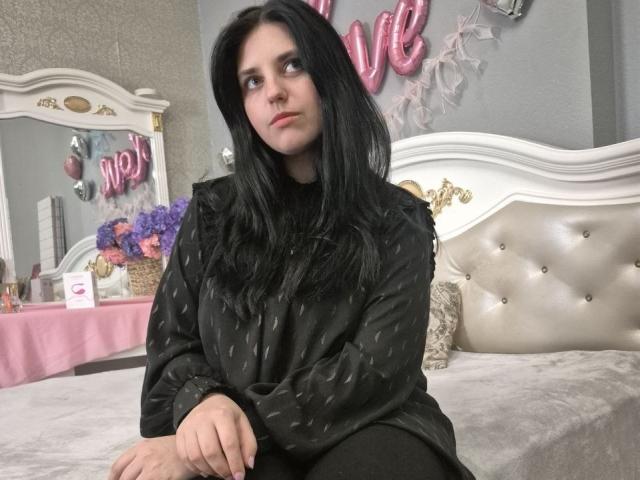 Esfil, con lunghi capelli neri, seduta su un letto bianco imbottito, indossa una camicetta nera con motivi e guarda verso l'alto con un'espressione leggermente espressione seria.
palloncino rosa con la scritta "AMORE" sullo sfondo. Decorazioni scintillanti..