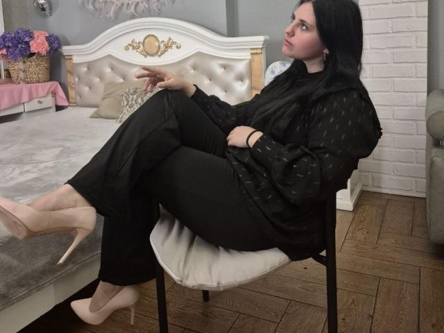 Esfil with black hair and pale skin, wearing a black dress and nude high heels, sitting on a white chair with legs crossed, showing off her toned legs and relaxed posture.
