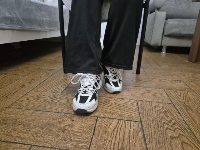 Esfils voeten, met zwarte en witte sneakers, staan op een houten parketvloer. Zwarte broek hangt los. Grijze bank en tafel op de achtergrond. Seksuele spanning door de nonchalante, licht provocerende pose..