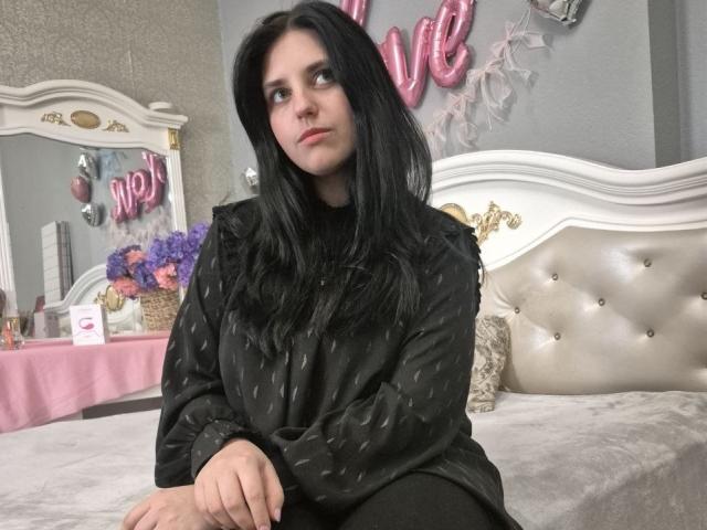 Esfil with long black hair, wearing a black polka-dot dress, sits on a tufted white bed. Pink balloon letters "LOVE" in the background. Sexual tension, casual bedroom setting.