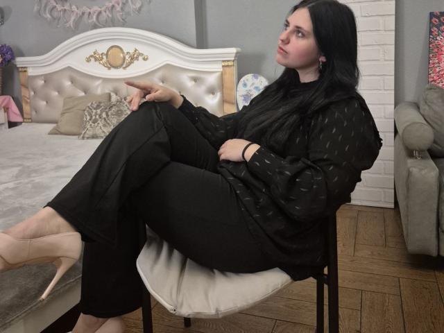 Esfil, con i suoi lunghi capelli neri, è seduta su una sedia bianca, con le gambe incrociate, indossando un vestito nero a pois, pantaloni neri e un paio di scarpe nude. tacchi alti, in una camera da letto con una testiera imbottita bianca..