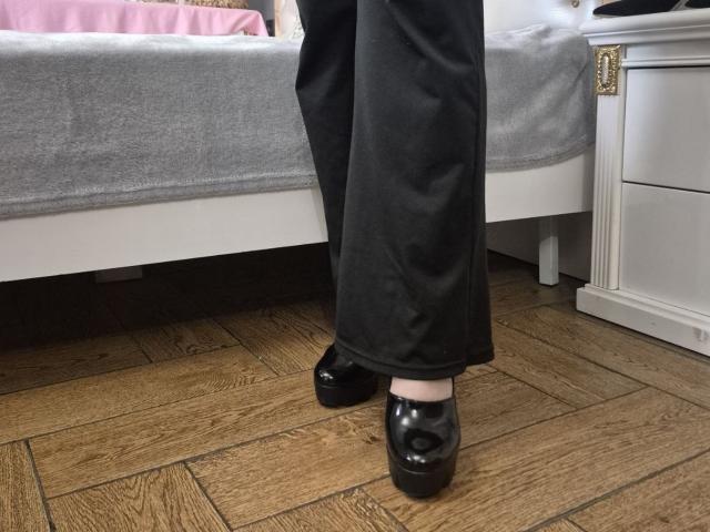 Esfil's lower legs and black loafers stand on wooden floorboards next to a white nightstand with gold details. Black pants, gray bed with white trim, soft lighting. Fucking sexy.