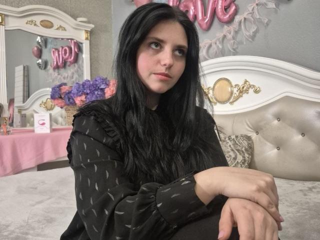 Esfil with long black hair sits on a tufted white bed, wearing a black polka-dot shirt, looking up with a slightly anxious expression. Pink balloons and a white mirror in the background.