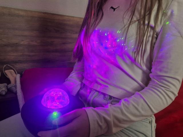 ZoieHurt sentada, con luces de neón moradas y azules brillando en sus manos, sudadera blanca con el logo de Puma, cabello castaño largo, de madera. Fondo de pared, sofá rojo, informal, íntimo, sexy, manos brillantes..