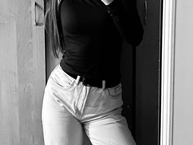 Black-and-white photo of ZoieHurt standing with her hips slightly cocked to the side. She's wearing a tight black top and high-waisted white jeans. Her long hair partially covers her face.