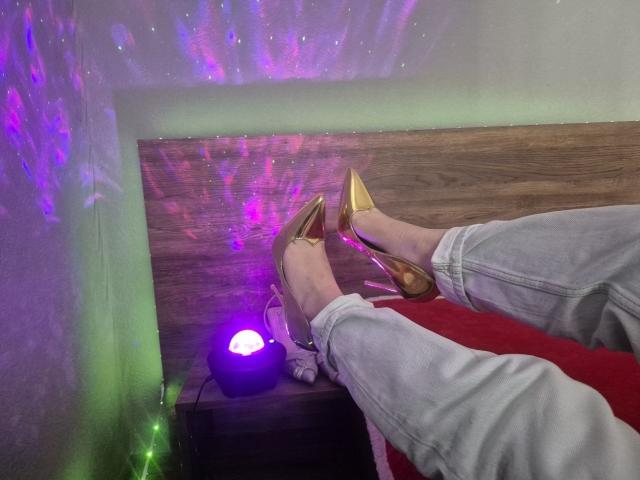 ZoieHurt's feet, clad in shiny gold high heels, rest on a wooden surface. She's wearing white jeans. Purple and green laser lights create a glowing effect in the background. Fucking sexy.