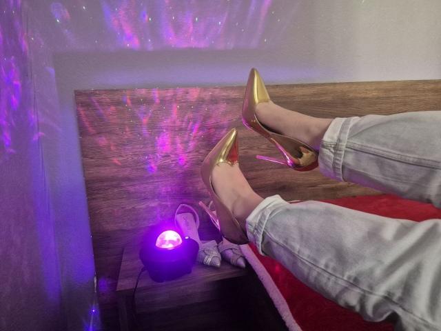 ZoieHurts Beine, die enge, graue Jeans und goldene High Heels tragen, ruhen auf einer Holzfläche. Lila und rosa Lichter werfen Reflexionen an die Wand und erzeugen eine verführerische Atmosphäre..