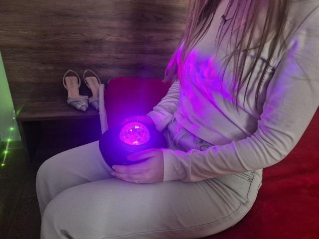 ZoieHurt sitting on a red chair, holding a glowing purple orb, wearing a beige hoodie and white pants. Her hands grip the orb suggestively, creating a charged, sexual atmosphere.