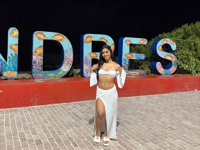 LaurentRay stands confidently in front of large colorful letters, wearing a white crop top and high-slit skirt, exposing her toned midriff and legs. Bright sunlight highlights her curvy figure.