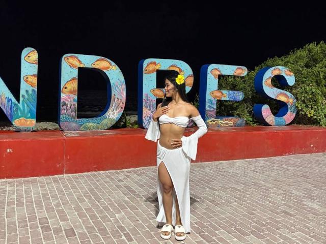 LaurentRay stands confidently in front of large colorful letters, wearing a white off-shoulder top and high-slitted skirt, showing off a toned midriff. Bright sunlight highlights her sexy, curvy figure.