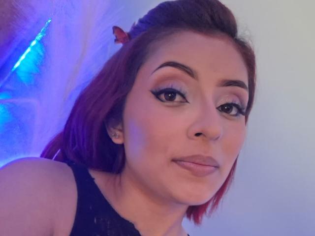 Nahaufnahme von LizBonnet, einer Frau mit hellbrauner Haut und roten Haaren, die schwarzen Eyeliner und ein schwarzes Oberteil trägt. Blaues Licht leuchtet im Hintergrund und erzeugt eine sexuelle Spannung..