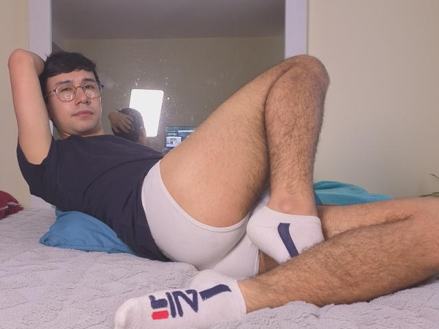 OliverHot69, un joven con gafas, camiseta negra, calzoncillos blancos, calcetines blancos con rayas negras y rojas, acostado en la cama. piernas separadas, pene visible, piernas peludas, pose casual, expresión relajada..