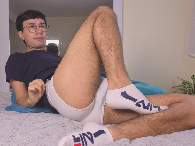 OliverHot69 está deitado em uma cama, com as pernas abertas, vestindo uma sunga branca com a inscrição "GU" e meias, com o pênis parcialmente visível. Ele usa óculos, tem pernas peludas, veste uma camiseta preta e há um travesseiro azul ao fundo. Pose casual e sensual.