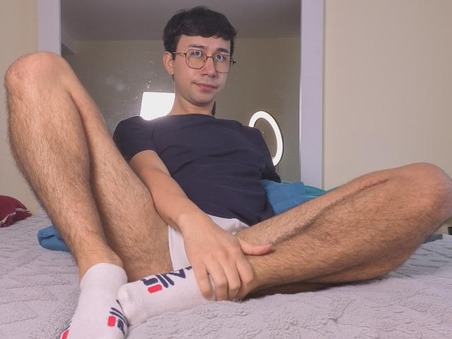 OliverHot69 sits on a bed, legs spread, wearing a black t-shirt and white socks with red and blue stripes. Hands on thighs, cock slightly visible, casual, nerdy, aroused. Soft lighting.