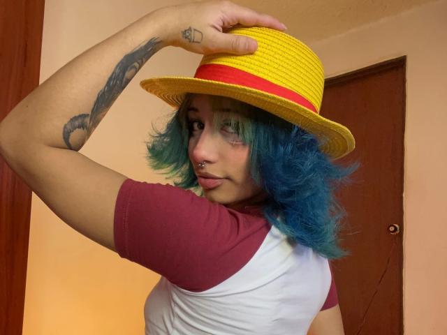 IsisJonnes69, blaue Haare, trägt einen gelben Strohhut mit rotem Band, ein dunkelrotes und weißes Hemd, tätowierter Arm, Nasenring. Hut justierend, sexy Pose, Innenaufnahme, Hintergrund: Holztür..