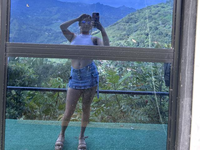 Saarah Jones è in piedi davanti a una finestra, tiene un telefono in mano, indossa un top corto bianco e pantaloncini di jeans blu, con una mano sulla sua testa. Montagne e vegetazione all'esterno. Riflessi sul vetro. Fa un caldo terribile..