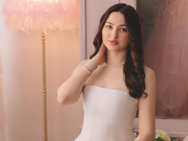 Photograph of ChristinaBliss with long, dark wavy hair, wearing a strapless white dress and pearl necklace, standing near a lit lamp, hand touching her neck, sexual tension, fuckable.