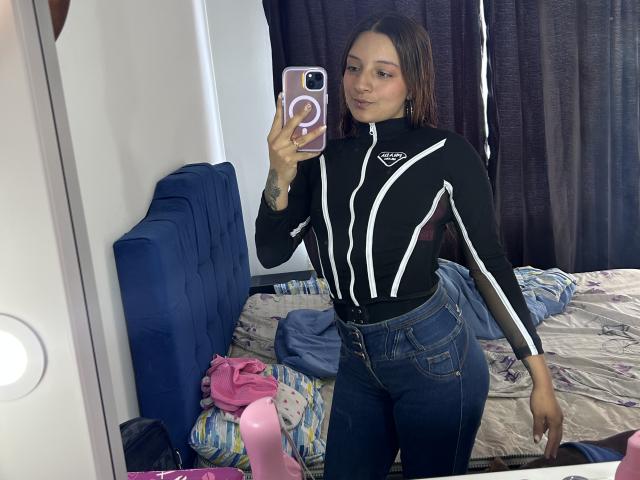 SaarahJones prend un selfie devant un miroir dans une chambre en désordre. Elle porte une combinaison noire avec des détails blancs et un jean taille haute. Ses cheveux mouillés collent à son visage. Incroyablement sexy. Gonflement visible..