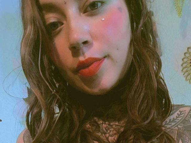 Foto em close-up de Amber Keller, com o rosto expressando tensão sexual. Ela tem cabelos castanhos ondulados, batom vermelho e um piercing no nariz. Seu ombro tatuado está parcialmente visível. Um olhar incrivelmente intenso..